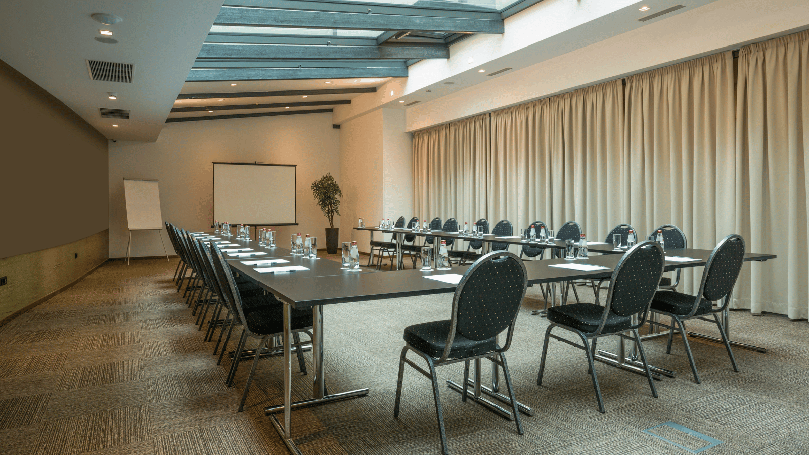 An empty conference centre with tables and chairs laid out in a u shape with a flip chart and projector screen at the front.