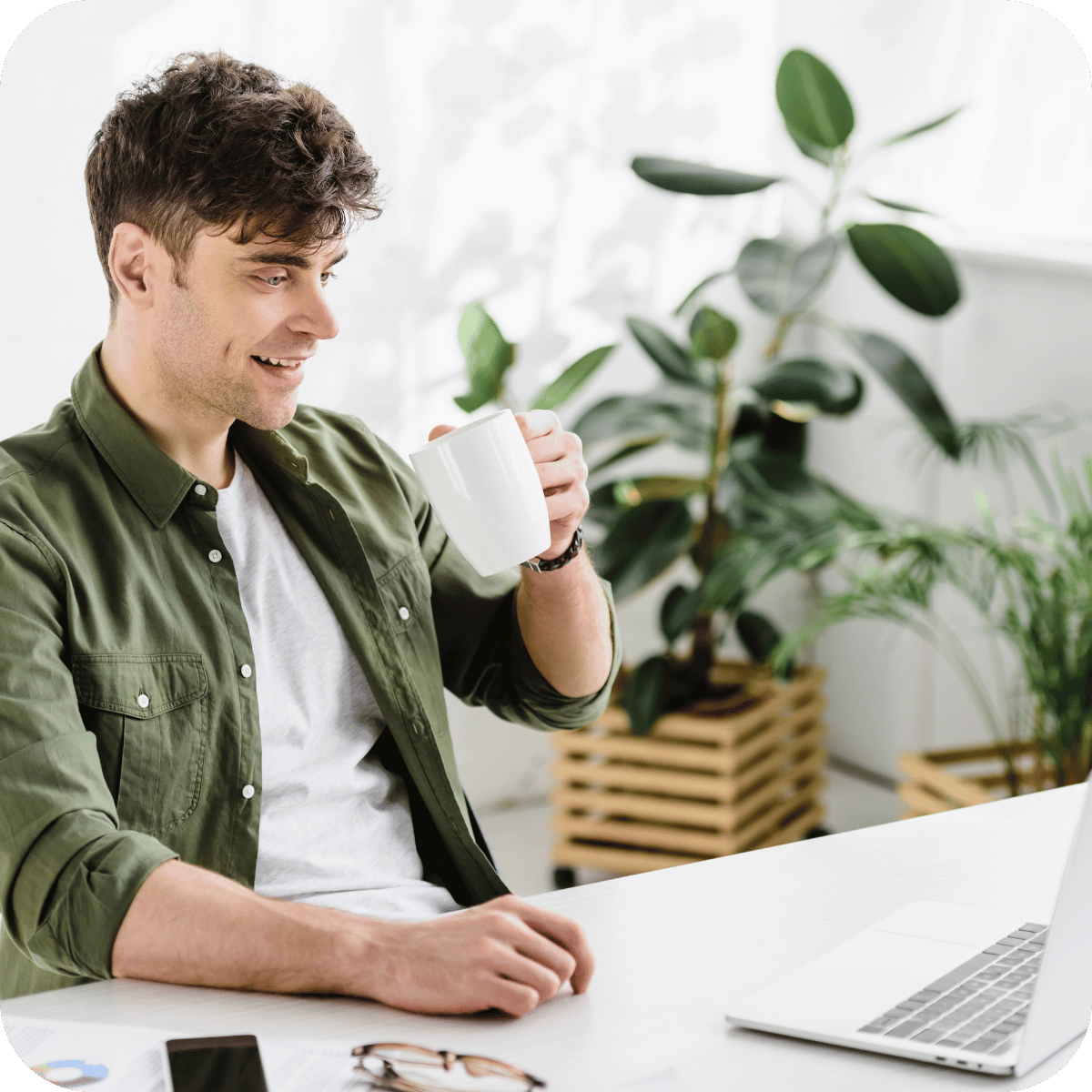 A man drinks a coffee in an office while sat at his desk on his laptop. 